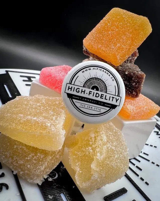 An assortment of sugar-dusted THC gummies in yellow, pink, and orange, stacked on a turntable with a High-Fidelity Cannabis Co. branded pick.