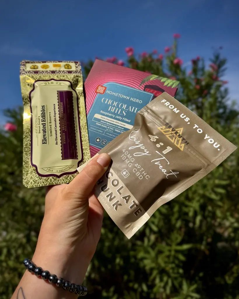 A hand holds three THC chocolate treats up against a green hedge and blue sky: Sweet Sensi Elevated Edibles orange creamsicle bar, Hometown Hero Chocolate Bites, and Seventh Hill 4:1 Crispy Treat.